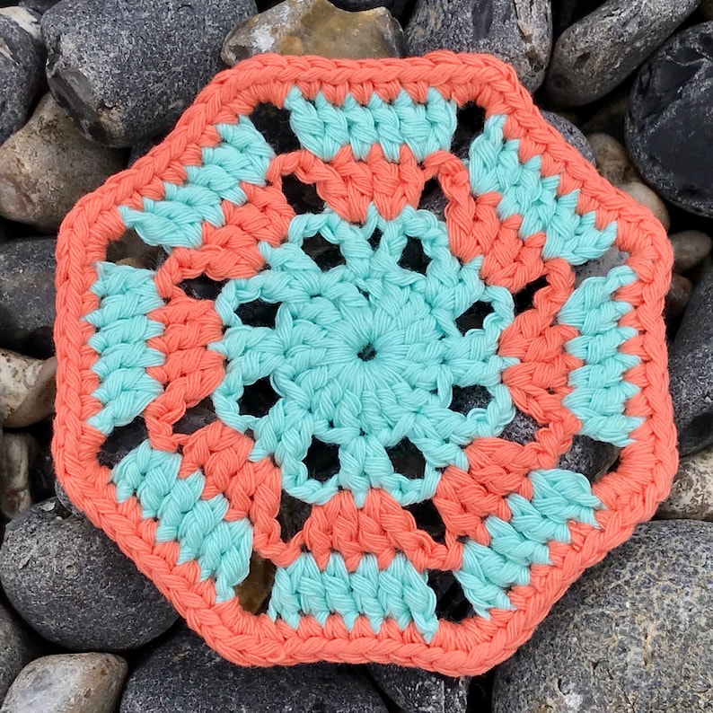 May include: A handmade, crocheted coaster in a hexagonal shape. The coaster features a central turquoise design surrounded by coral-colored sections, with a coral border. The coaster is placed on a bed of gray rocks.