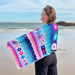 Puede incluir: Un chal de ganchillo hecho a mano y colorido con un diseño floral y geométrico. El chal presenta una vibrante mezcla de tonos rosa, morado, azul y turquesa. El diseño calado y los motivos florales crean una estética bohemia.