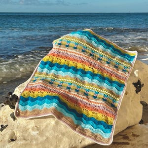 May include: A vibrant, crocheted blanket featuring a wave-like design in blue, yellow, orange, and pink hues. The blanket is displayed on a rock by the sea, highlighting its detailed pattern and hand-made quality. The blanket measures approximately 100 cm by 100 cm.