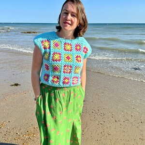 May include: A woman wearing a light blue crochet top with a pink, yellow and green geometric pattern. She is standing on a sandy beach with the ocean in the background. She is wearing a green skirt with a floral pattern.