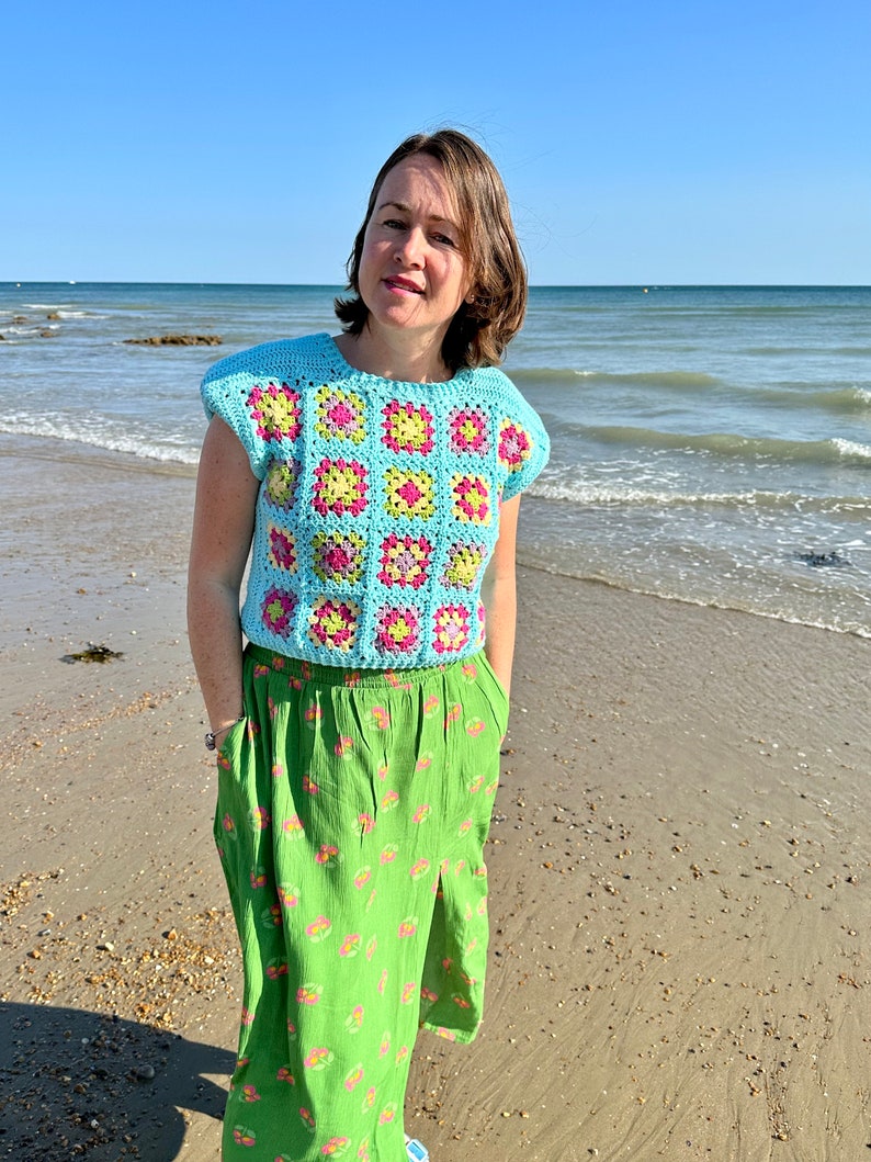 May include: A woman wearing a light blue crochet top with a pink, yellow and green geometric pattern. She is standing on a sandy beach with the ocean in the background. She is wearing a green skirt with a floral pattern.