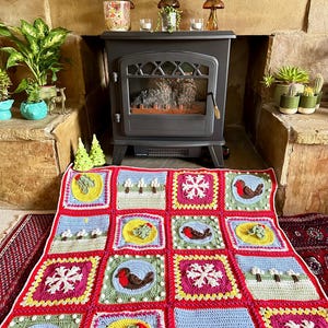 Puede incluir: Una manta de ganchillo colorida con un tema navide&ntilde;o. La manta est&aacute; hecha de cuadrados con diferentes dise&ntilde;os, incluyendo copos de nieve, petirrojos, acebo y &aacute;rboles. La manta est&aacute; colocada en el suelo frente a una chimenea.