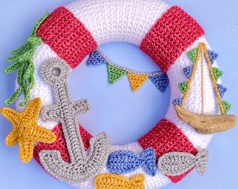 May include: A crocheted wreath in the shape of a life preserver with red and white stripes. The wreath is decorated with crocheted nautical themed items, including a sailboat, an anchor, a starfish, and fish. There are also small crocheted flags in green, yellow, and orange.