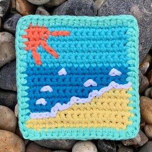 May include: Handmade crochet coaster featuring a beach scene. The design includes a yellow beach, blue ocean, white waves, and a coral-colored sun. The coaster is square with a light green border, perfect for adding a touch of summer to any space.