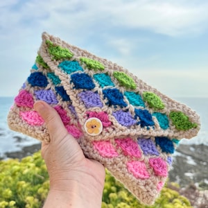 Pode incluir: Uma carteira de mão de crochê com um padrão geométrico colorido em tons de verde, azul, roxo e rosa. A carteira tem um fecho de botão com um desenho de flor amarela.