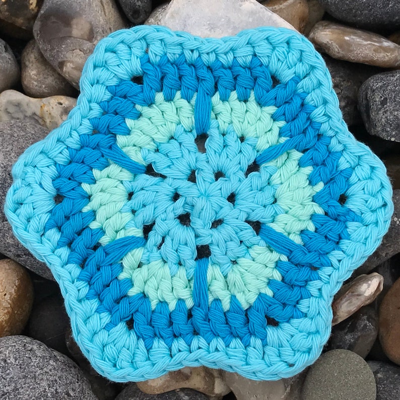 May include: A handmade, crocheted coaster in a six-pointed star shape. The coaster features a color palette of light blue, medium blue, and teal. The coaster is made of cotton yarn and is suitable for use with hot or cold beverages.