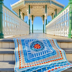 May include: A colourful crocheted blanket with a geometric pattern in shades of blue, red, yellow, and white. The blanket is lying on the steps of a white and green gazebo with a white railing.