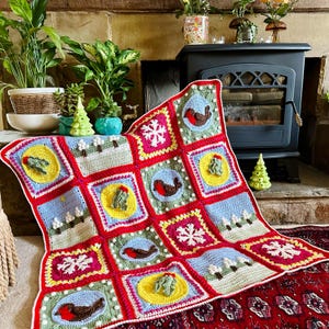 Puede incluir: Una manta de ganchillo colorida con un tema invernal. La manta presenta cuadrados con diferentes dise&ntilde;os, incluyendo petirrojos, copos de nieve y &aacute;rboles. La manta est&aacute; colocada sobre una alfombra delante de una chimenea.