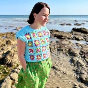 May include: A woman wearing a light blue crochet top with a colourful granny square pattern. The top has short sleeves and a round neckline. She is standing on a beach with rocks in the background.