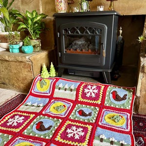 Puede incluir: Una manta de ganchillo colorida con un tema navide&ntilde;o. La manta presenta cuadrados con diferentes dise&ntilde;os, incluyendo copos de nieve, petirrojos y acebo.