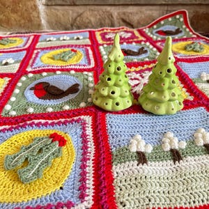 Puede incluir: Una manta de Navidad de ganchillo con un esquema de color rojo, blanco y verde. La manta presenta varios motivos navide&ntilde;os, incluyendo un petirrojo, acebo y una escena de invierno. Dos peque&ntilde;os &aacute;rboles de Navidad de cer&aacute;mica verde se colocan encima de la manta.