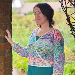 Op de afbeelding: Een kleurrijk, wikkel-gehaakt vest met lange mouwen. Het vest heeft een mix van pastelkleuren, waaronder blauw, paars, roze en groen. Het vest wordt gedragen over een teal groene jurk. De achtergrond omvat een bakstenen muur en groen.