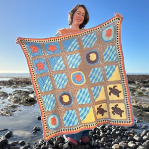 Puede incluir: Una manta de ganchillo hecha a mano con un diseño de temática marina. La manta presenta cuadrados con patrones de olas azules, estrellas de mar, tortugas y motivos de conchas marinas en tonos azules, marrones, naranjas y amarillos. La manta se sostiene contra un cielo azul claro.