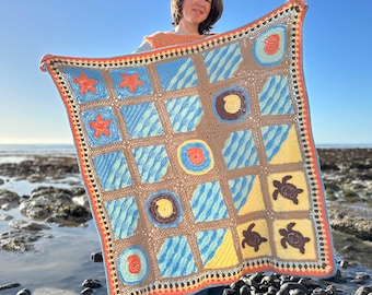 May include: A handmade crochet blanket featuring a sea-themed design. The blanket showcases squares with blue wave patterns, starfish, turtles, and seashell motifs in shades of blue, brown, orange, and yellow. The blanket is held up against a clear blue sky.