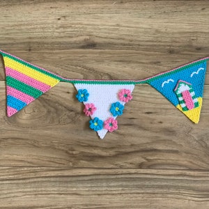 Könnte beinhalten: Eine gehäkelte Girlande mit Strandthema. Die Girlande hat drei Flaggen, eine mit Streifen in Pink, Grün und Gelb, eine mit weißem Stoff und rosa, blauen und weißen Blumen und eine mit einer blauen und grünen Strandhütte mit Möwen.