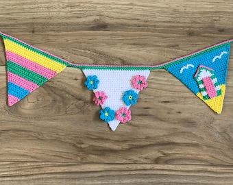 May include: A crocheted bunting with a beach theme. The bunting has three flags, one with stripes of pink, green, and yellow, one with white fabric and pink, blue, and white flowers, and one with a blue and green beach hut with seagulls.