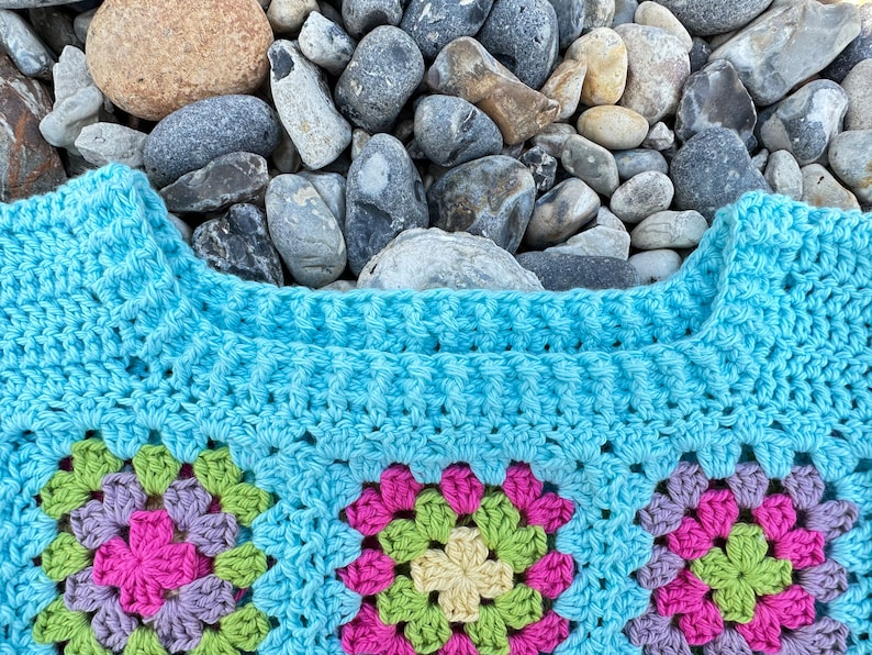 May include: A blue crocheted granny square blanket with a colourful floral pattern. The blanket is made with a variety of different coloured yarns, including pink, green, yellow, and purple.