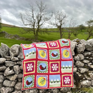 May include: A colourful crocheted blanket with a winter theme. The blanket is made up of squares with different designs, including snowflakes, robins, and trees. The blanket is draped over a stone wall.