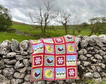 May include: A colorful crocheted blanket with a winter theme. The blanket is made up of squares with different designs, including snowflakes, robins, and trees. The blanket is draped over a stone wall.