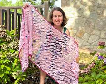 May include: A pink crocheted shawl with a delicate floral pattern. The shawl has a burgundy border and is being held by a person in a garden setting.