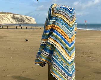 May include: A crocheted blanket with a blue, green, yellow, and white color scheme. The blanket is draped over a wooden post on a beach.
