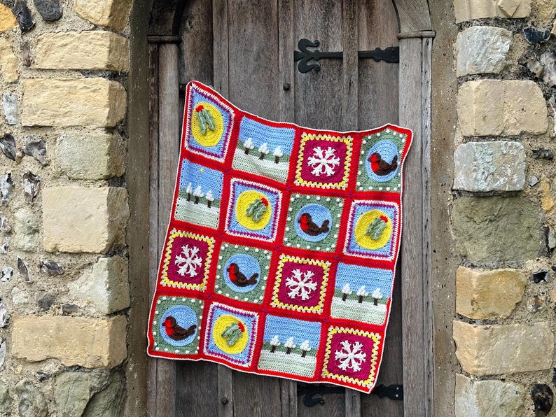 Puede incluir: Una manta de crochet roja, verde, azul y amarilla con tem&aacute;tica navide&ntilde;a. La manta est&aacute; compuesta por cuadrados con diferentes dise&ntilde;os, incluyendo copos de nieve, petirrojos, acebo y &aacute;rboles.