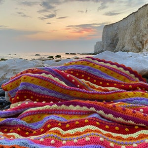 May include: A colourful crocheted blanket with stripes of orange, yellow, pink, purple, and white. The blanket is laid out on a rocky beach with a white cliff in the background.