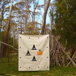 May include: A cream-coloured Halloween-themed quilt featuring three witch hats and a pumpkin. The quilt is decorated with black star shapes and has a black border. The quilt is displayed outdoors in a forest setting.