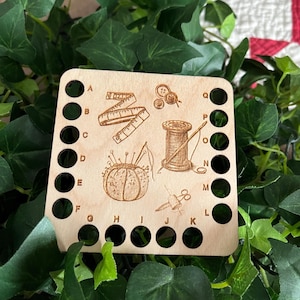 May include: A wooden sewing organizer with engraved illustrations of sewing tools, including a measuring tape, buttons, a pincushion, thread spool, needle, and scissors. The organizer has multiple holes for thread storage.