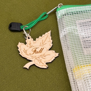 May include: A light-colored wooden maple leaf charm featuring the engraved word Fall in a script font. The charm is attached by a silver-toned metal lobster clasp to a green braided cord, which is connected to a clear, textured plastic pouch.