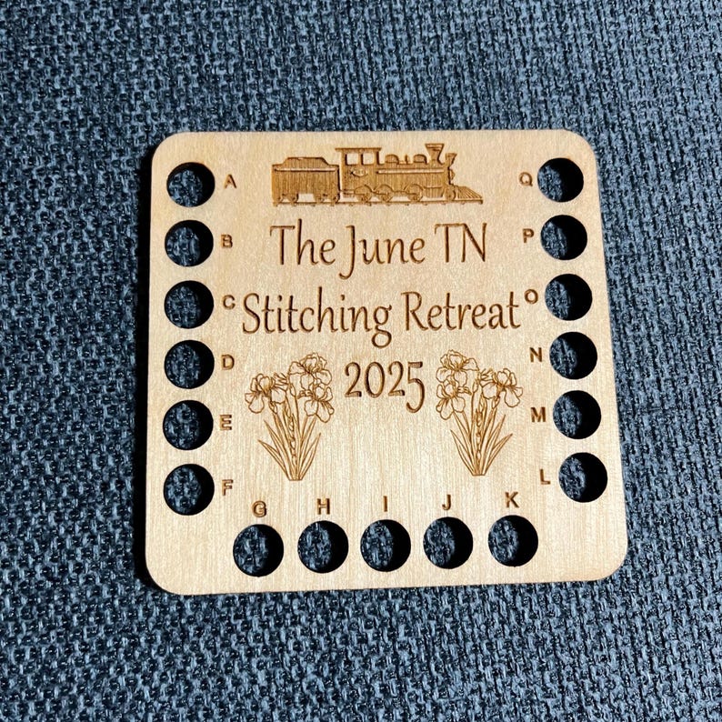 May include: A wooden needle minder with a train illustration and the text "The June TN Stitching Retreat 2025." It has multiple holes for organizing threads, and floral designs. The needle minder is light brown.