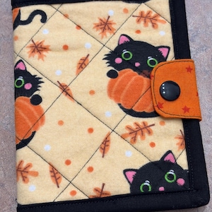 May include: A quilted, rectangular cover with a black border. The cover features a light yellow background with a repeating pattern of black cats holding orange pumpkins, orange leaves, and white dots. An orange tab with a black button secures the cover.