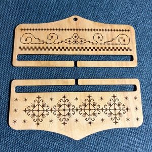 May include: Two wooden weaving tools with decorative etched designs. The top tool has a series of dots and a zigzag pattern. The bottom tool features a symmetrical floral design with star accents. Both tools are light brown.