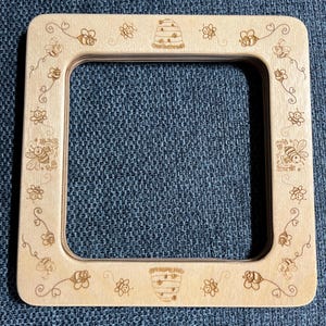 May include: A square light-colored wooden frame featuring laser-engraved designs of bees, flowers, and beehives along all four borders. The frame has rounded corners and a smooth, natural wood grain finish with a hollow center opening.