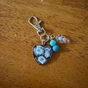 May include: Silver-toned keychain with a heart-shaped pendant featuring blue floral design. The keychain includes turquoise beads and a clear flower-shaped charm. The background is a wooden surface.