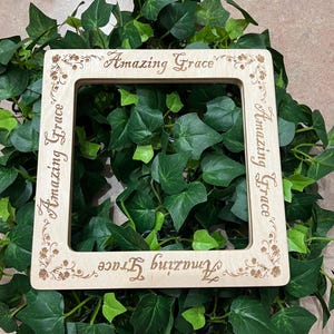 May include: A square light-colored wooden picture frame with rounded corners. The frame features the laser-engraved text "Amazing Grace" on all four sides, accompanied by small floral vine motifs etched into the wood at each corner.