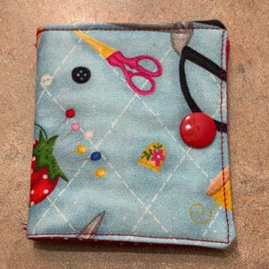 May include: A square, fabric sewing kit with a light blue background and a red border. The kit features a pattern of sewing tools, including scissors, needles, and a strawberry. A large red button secures the kit closed.