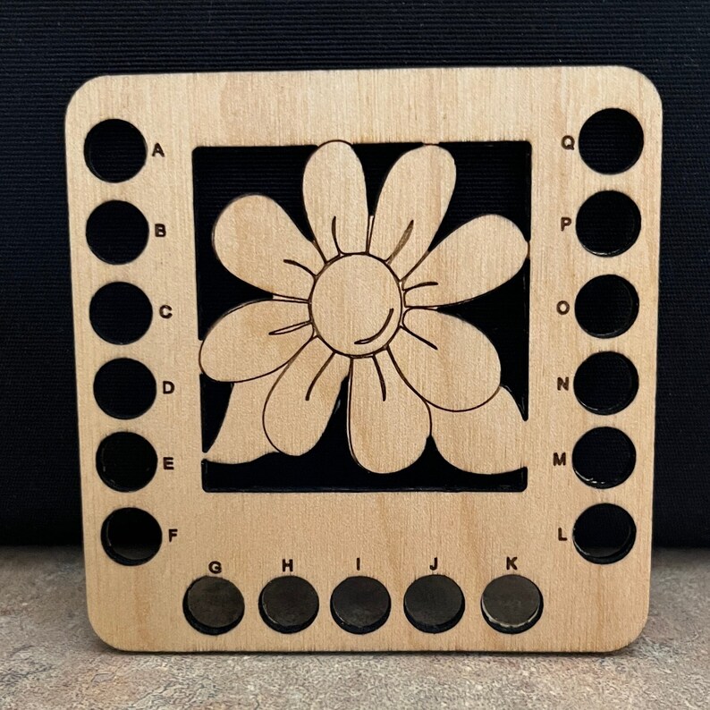 May include: Wooden yarn bowl with a flower design and 17 holes for holding yarn. The holes are labeled with letters from A to Q.