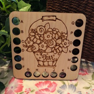 May include: Wooden stitch holder with 18 holes for holding yarn. The holder is decorated with a floral design and the word "fleur".