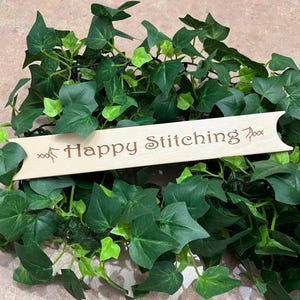 May include: A wooden sign with the text "Happy Stitching" in brown lettering, resting on a bed of green ivy. The sign has a light wood color and a decorative edge. The ivy has dark green leaves.