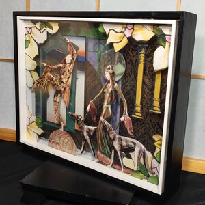 May include: A framed mixed media artwork featuring two figures in a stylized setting. The figures are dressed in elaborate costumes and are surrounded by geometric shapes and floral patterns. The artwork is set against a backdrop of a patterned wallpaper.