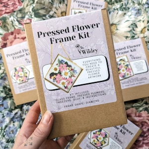 May include: A pressed flower frame kit with a diamond-shaped frame. The kit includes a glass frame, pressed flowers, tweezers, glue, and instructions. The frame is gold and has a chain attached to it. The flowers are pink, white, and yellow.