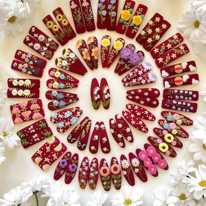 May include: A collection of 30 red velvet hair clips with embroidered floral designs. Each clip features a different floral pattern, including sunflowers, daisies, and lilies.