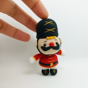 May include: A crocheted toy soldier with a black hat, red uniform, and a yellow belt. The toy has a white beard, black mustache, and a red nose.