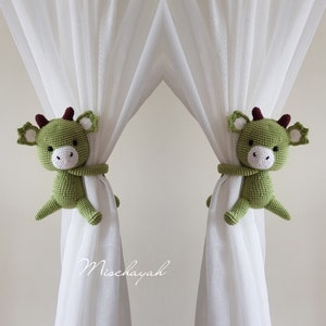 May include: Two green crocheted dragon curtain tiebacks with white eyes and red horns. The tiebacks are holding back white sheer curtains.