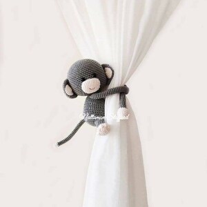 May include: A gray crocheted monkey toy holding a white curtain. The monkey has a pink nose and a happy expression.