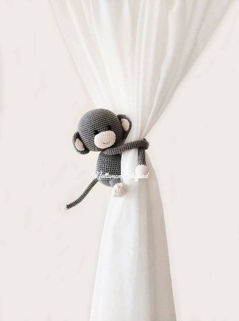 May include: A gray crocheted monkey toy holding a white curtain. The monkey has a pink nose and a happy expression.