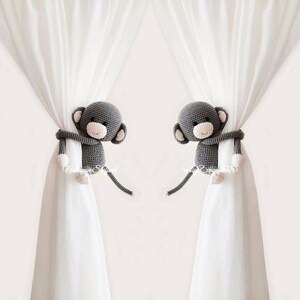 May include: Two gray crocheted monkey curtain tie backs. Each monkey has a white face with black eyes and a pink nose. The monkeys are holding onto white curtains.