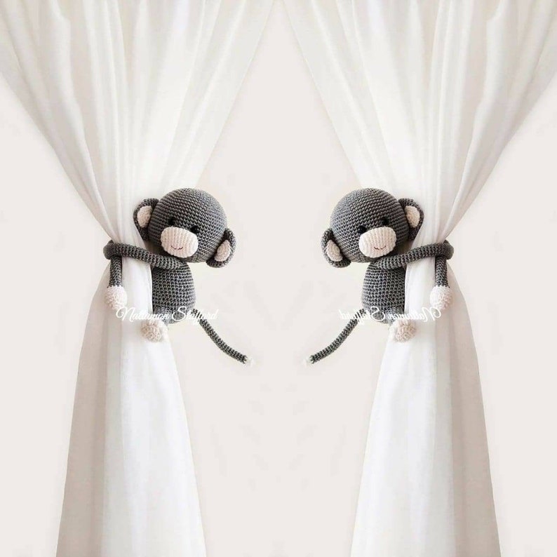 May include: Two gray crocheted monkey curtain tie backs. Each monkey has a white face with black eyes and a pink nose. The monkeys are holding onto white curtains.