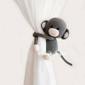 May include: A gray crocheted monkey toy with pink accents is used as a curtain tieback. The monkey is holding the curtain with its arms and has a long tail.